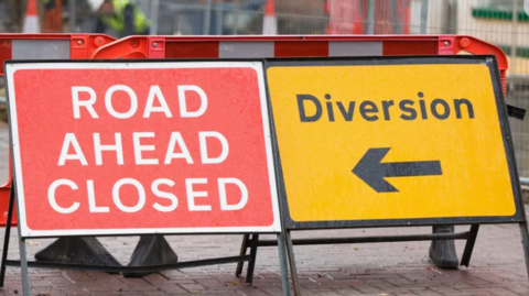 A road ahead closed sign, with red background and white writing. And a diversion sign with an arrow pointing left, in black writing on a yellow background. There are traffic cones in the background.