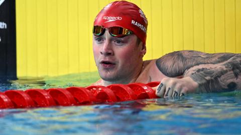 Adam Ramsay-Peaty, wearing a red Speedo swimming cap and goggles, holds the red lane separator in the pool after the 50m breaststroke heats at the 2026 Commonwealth Games