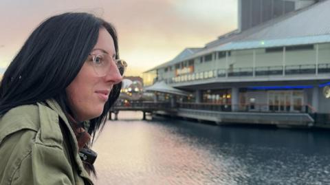 A lady looking across water, the sky is light pink and she has black hair. She's wearing clear glasses and a green coat. 