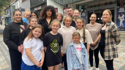 A group of adults and children stand together on a paved street in front of a set of automatic glass doors