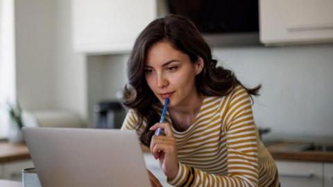 A generic photo of a woman in a striped brown and white top looking at a laptop. Cupboards are blurred behind her.