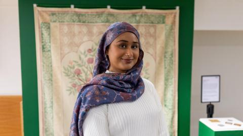 A woman wearing a purple floral headscarf and white jumper smiling at the camera. Behind her is a tapestry with pink flowers and green leaves. 