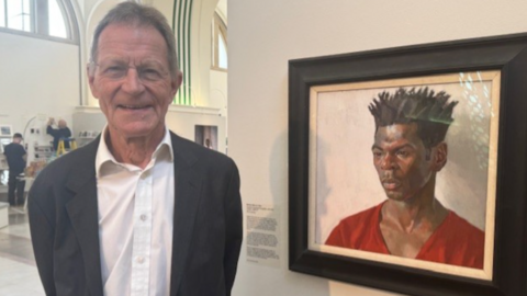 Wearing a grey a suit over an open neck white shirt, a smiling Sir Nicholas Serota is also wearing a pair of round, almost rimless glasses. He's standing alongside a portrait of the artist Nahem Shoa.