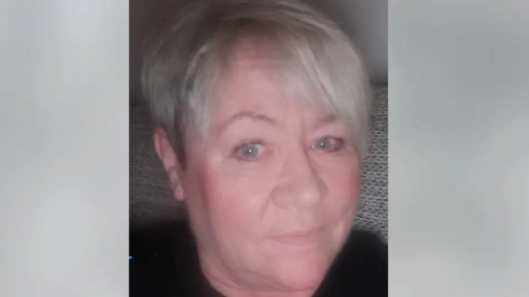 Kelly Faiers is looking ahead. She has silvery grey hair and is wearing a black top.