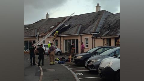 Firefighters are using a long ladder to access the roof of a block of flats and damage can bee seen to roofing tiles.