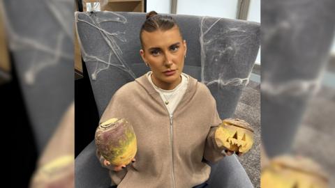 A woman sat on a grey chair in a beige cardigan. She is holding two turnips.