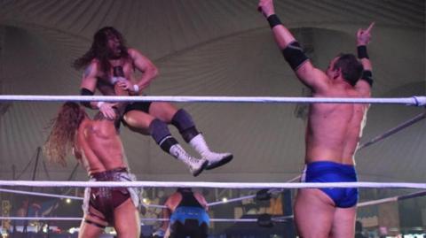 Four wrestlers inside a ring. One with his back to us is lifting another in the air around the throat and is about to slam him down. Another man is on the right with both hands raised in the air. A fourth man is slumped over the ropes on the far side. 
