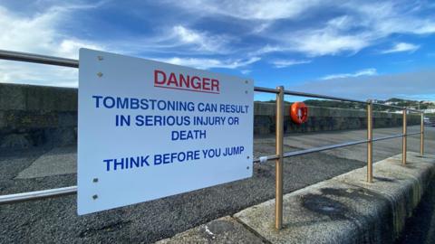 A sign attached to a railing near a sea wall that reads: Danger, tombstoning can result in serious injury or death, think before you jump. In the background there is a life ring attached to the sea wall.