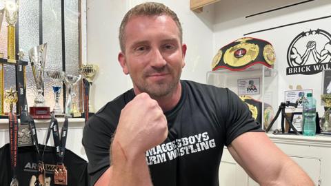 James "Brick" Wall. He is showing his right fist to the camera. To the left are various trophies which he has won. He is wearing a black T-shirt with the wording Maggie's Boys Arm Wrestling. 