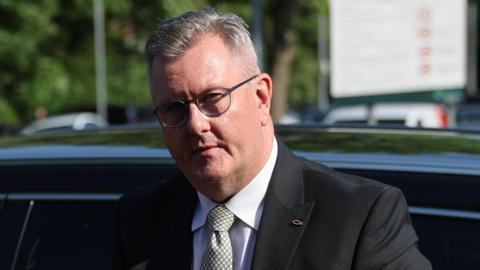 Sir Jeffrey Donaldson has short, cropped grey hair. He is wearing black-rimmed glasses and a dark grey suit, white shirt and grey patterned tie. He is standing in front of a black car and is carrying a brown leather satchel bag in front of him.