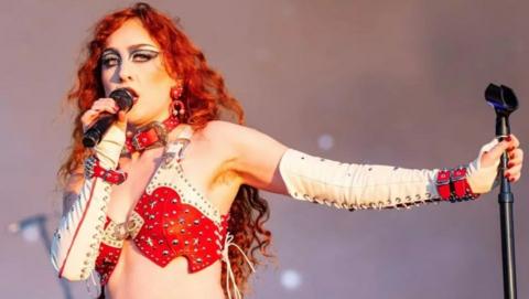 Chappell Roan performing on stage with thick eyeliner and heavy eye makeup, long red curled hair and a red and white outfit. She is singing into a microphone. 