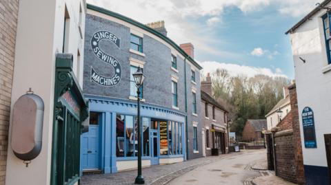 A curved street with Victorian buildings on both sides. one with a brightly painted blue front and the words "Singer Sewing Machines" on the wall.
