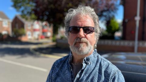 A man with grey hair and a short beard and moustache with black sunglasses is wearing a blue shirt and looking at the camera with a serious expression. The background is blurred and there is a road with houses in sight.