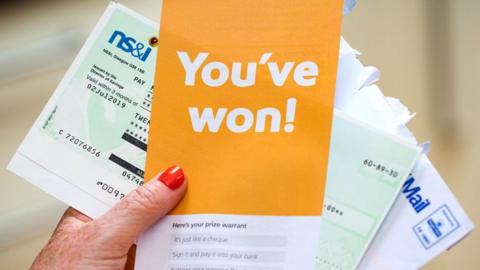 A person's hands holding Premium Bonds literature, including a leaflet saying they have won a prize and a cheque for £25. 