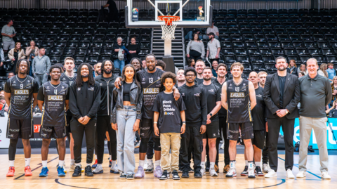 Darius Defoe, a man in his 40s who is six feet and eight inches tall, towering in the middle of a basketball court amongst the Newcastle Eagles basketball team and members of his family.