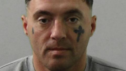 Mugshot of Wood. He is clean-shaven and has a large cross tattooed under his left eye and a teardrop to the left of his right eye.