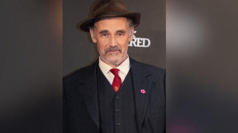 Sir Mark Rylance smiles into the camera wearing a navy pinstripe suit and waistcoat with a red tie and a brown fedora hat.