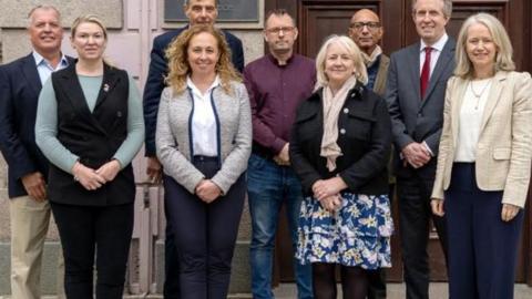 Nine people stood in two rows consisting of five men at the back and four women in front of them stood all wearing smart casual clothing stood outside a building looking towards the camera.