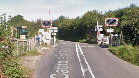 The level crossing on Cathole Bridge Road. There are green fields to the left of the carriageway. The road joins a track in the distance. There is shrubbery to both sides of the carriageway.