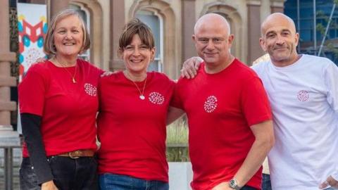 Two women stand in a line next to two men. The two women and one of the men all wear the same red T-shirts. The other man on the end wears a white long sleeved T-shirt. One of the women has bobbed blonde hair while the other has brown hair that has been tied up. Both men are bald. 