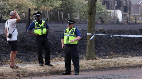 Police officers talk to a man at a cordon in Stourbridge. Behind them a grass field is now black after being burned by fire