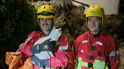 Firefighters Sam Hunt (left) and Dom Lever (right) holding a white cat. The men are wearing yellow helmets and red firefighter gear. 