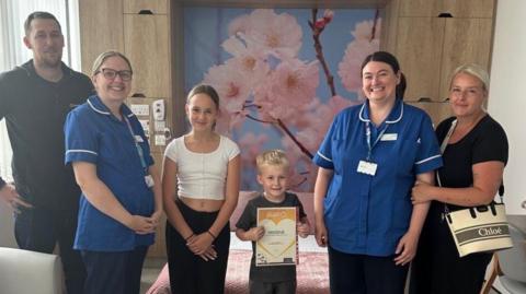 Two nurses in blue NHS uniforms, a tall brown-haired man, blonde woman, young teenage girl and small boy stand in a line smiling at the camera.