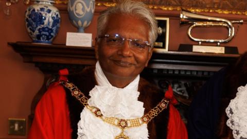 Wearing his mayoral robes and gold chain, Bharat Pankhania smiles at the camera. Behind him two vases and a musical instrument can be seen on a mantelpiece.