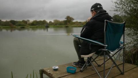 A young person is seen from the side sitting on a camping chair which is placed on a wooden jetty. They are holding a fishing rod out into a large lake in front of them and there is a tub of bait on the floor next to them