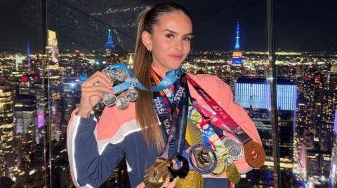 Lucy Scott has eight medals hanging from her neck. They vary from gold, silver and bronze with pink and blue coloured ribbons. She is stood in front of the view of New York City lit up at night. 