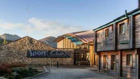 A wooden building sits to the left of the frame. A stone wall which reads 'Tollymore National Outdoor Centre' sits to the left of the building. Behind the building is are two large mountain peaks which split the light blue sky. 
