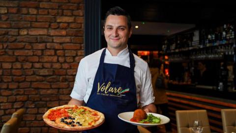 Jason Maimone smiles as he holds a pizza and another Italian dish in his restuarant. He wears a blue apron with the Italian tricolour over his chef whites.