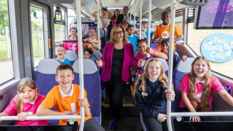 West of England mayor Helen Godwin stood in the aisle of a bus. There are several children sat on the bus seats. All of them are looking at the camera and smiling.