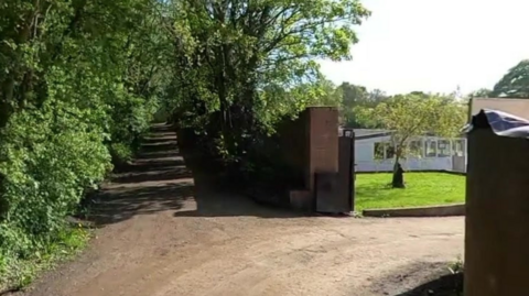 Offerton Lane - wide track surrounded by trees, which splits off to the right where there is an open gate and a white building behind a patch of vibrant green grass.