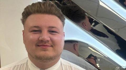 Picture of Ryan Featherstone smiling at the camera wearing a white shirt. Ryan has mousey blond brushed-back hair and bearded stubble