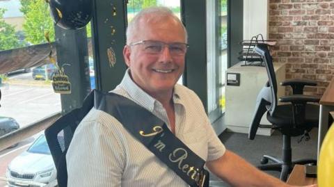 A man in his 60s sits at a desk with balloons and a sash marking his retirement