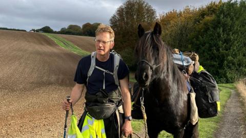 Roger Sewill during a trek in a field next to Scarlet. She is carrying his bags.
