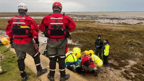 Emergency crews huddle around the casualty on marshy land. A sloping wall leads up to a higher vantage point in the foreground, where more service members look on.