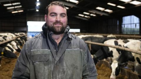 Gethin Hughes, dairy farmer stands in his cattle shed wearing blue overalls
