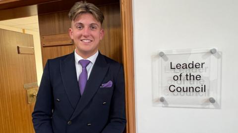 George Finch dressed in a navy suit and purple tie, stood outside an office with a sign leader of the council