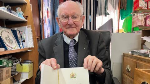 Arthur Ferguson in his haberdashery. He is wearing a black suit with a jumper underneath. He has grey hair and bald on top. He has thin-rimmed glasses and a tie with a white spotted pattern. He is holding his letter from the King and Queen, which he received when he turned 100.