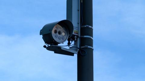 A  small black camera attached to a black pole. Behind it is a clear blue sky.