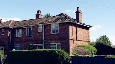 The fire-damaged house. There are damaged tiles on the roof and burn marks around the windows and door.