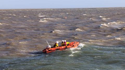 Three RNLI volunteers are sailing in an orange inflatable rescue boat in choppy waters. They are wearing yellow and red uniforms and white helmets. 