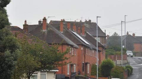 A street with red brick houses, there is grey sky in the background and the pavement runs along the right-hand side of the image, with a silver car parked in the corner. There are also small green bushes in front of the house. 