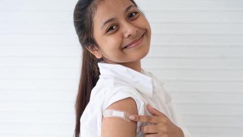 A girl points to a sticking plaster on her upper arm, after being vaccinated. She is smiling at the camera and wears a white school shirt and has her hair tied back in a ponytail. 