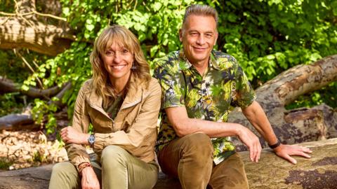 Michaela Strachan and Chris Packham, sitting on a log in front of a green bush