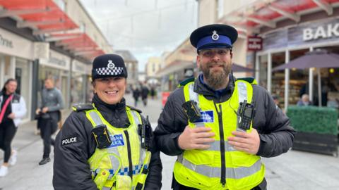 PC Jill Robertson and PCSO Dan Snell on patrol in Workington town centre. They are both in uniform, with hats and high-viz vests. They are standing next to each other and smiling at the camera. PC Robertson's hair is tied back. PCSO Snell has a beard and septum piercing.