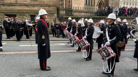 Members of a Royal Navy band in full uniform with white helmets and carrying drums march past a church building with the public looking on