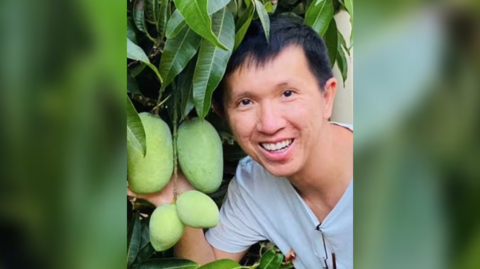 Thanh Phan smiling next to a mango tree with immature fruit